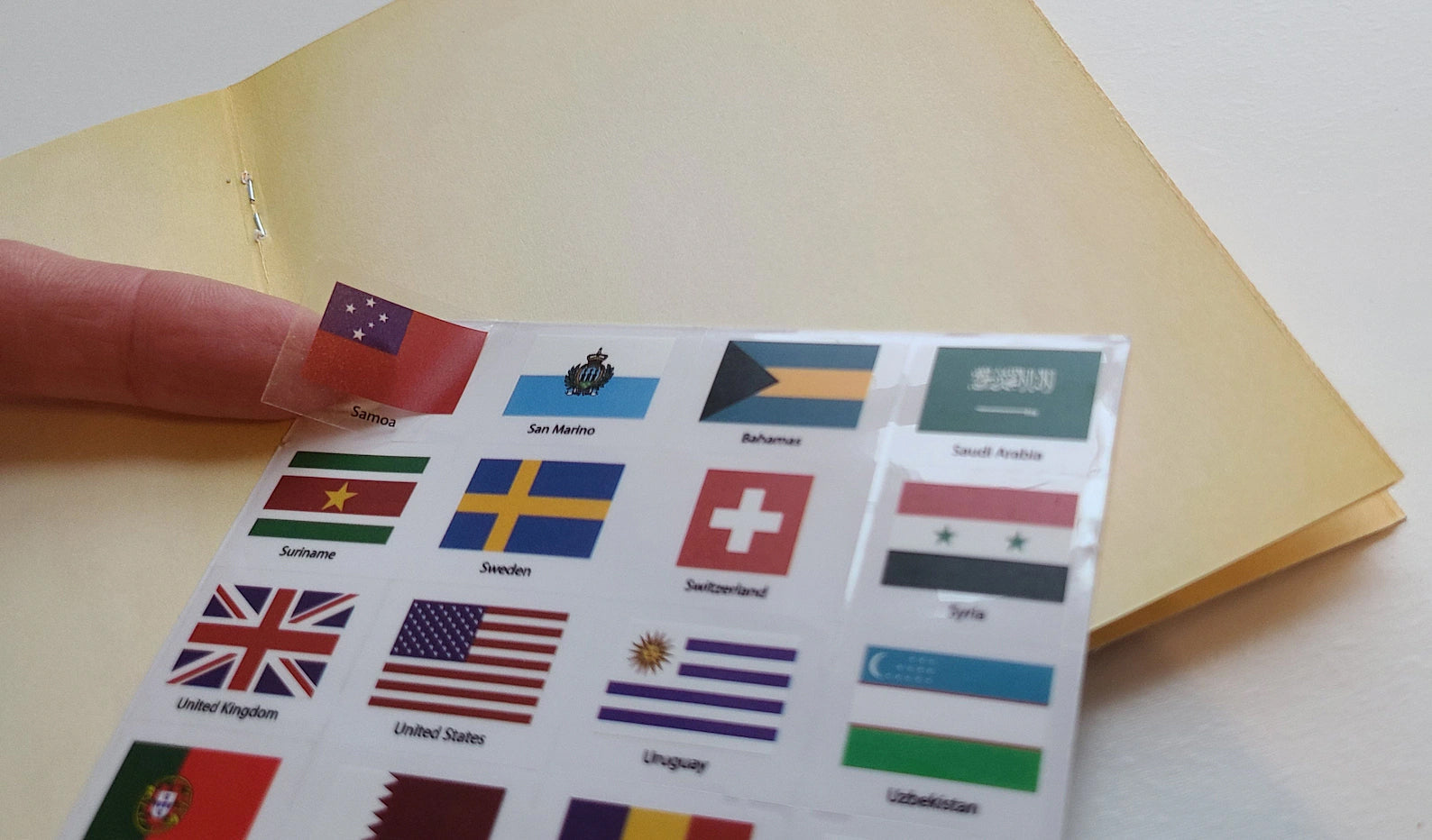 Hand peeling a world flag sticker from a sheet of 192 country flags — passport booklet in background, world flag sticker set by SJPrinter, Cherry Hill NJ